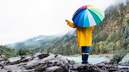 A person stands with a colorful umbrella near a mountain lake. 